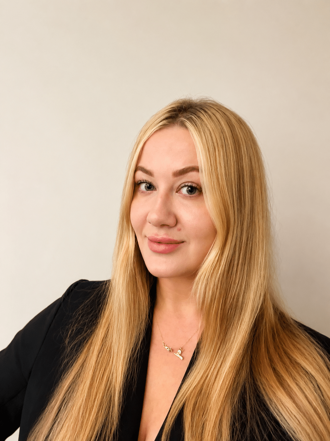 A confident woman with long blonde hair poses in a black blazer against a neutral background,