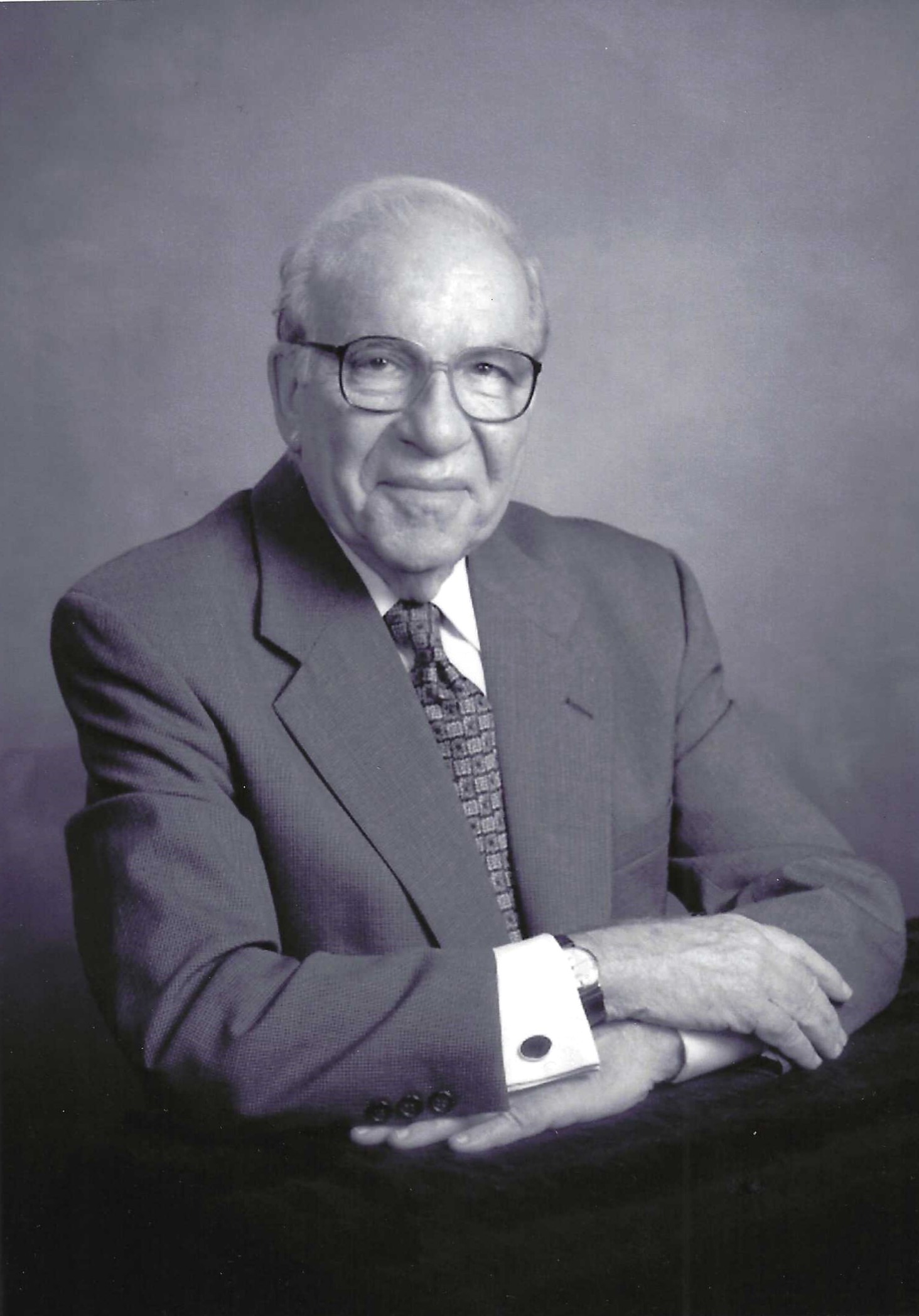 Elderly man in a gray suit, glasses, and patterned tie, smiling confidently with arms crossed
