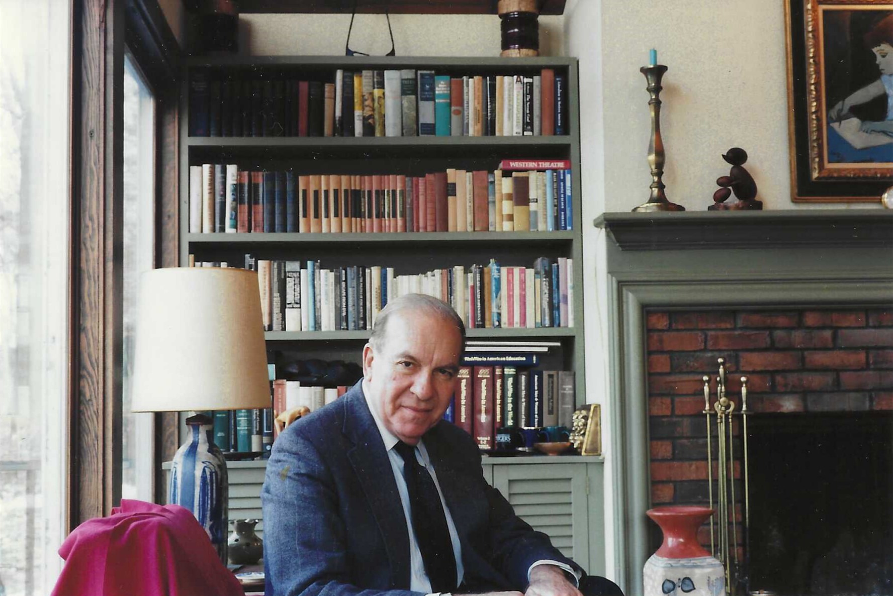 An older man in a suit sits at a table in a cozy room,