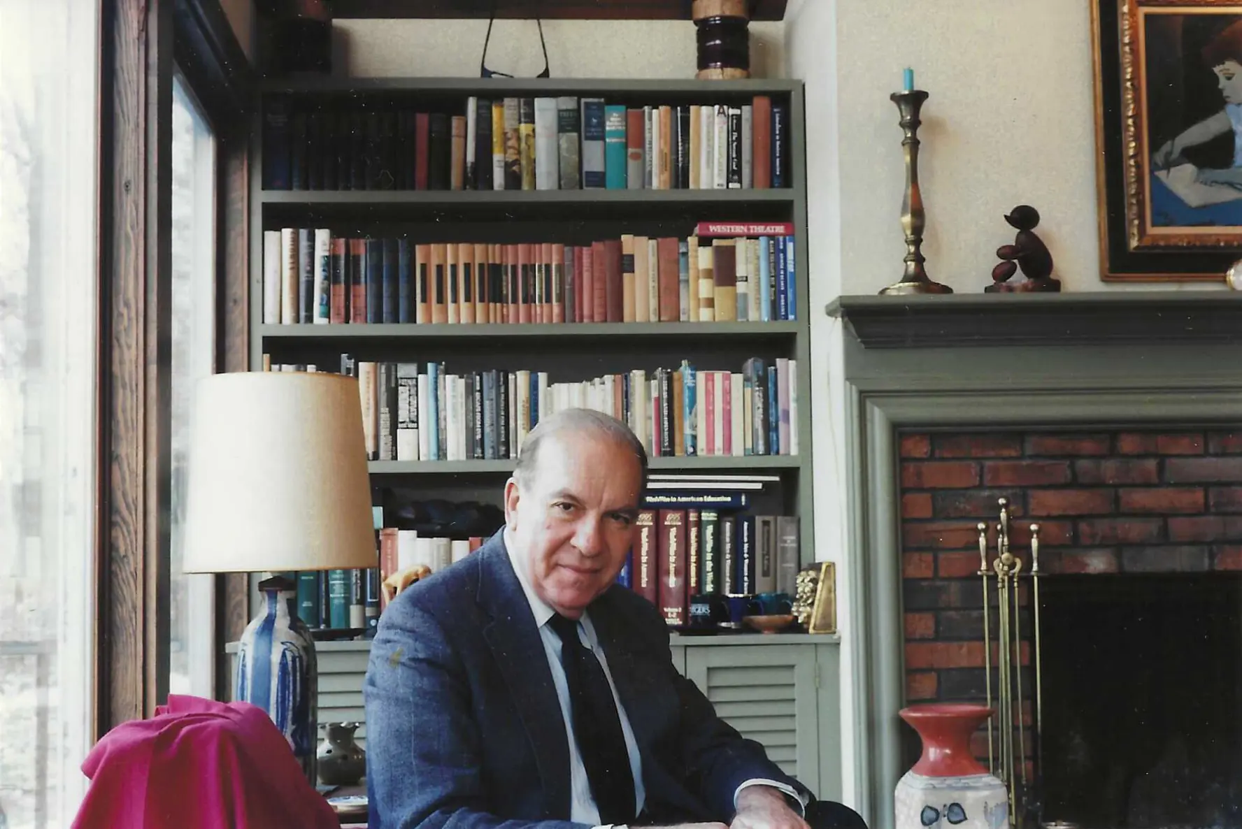 An older man in a suit sits at a table in a cozy room,