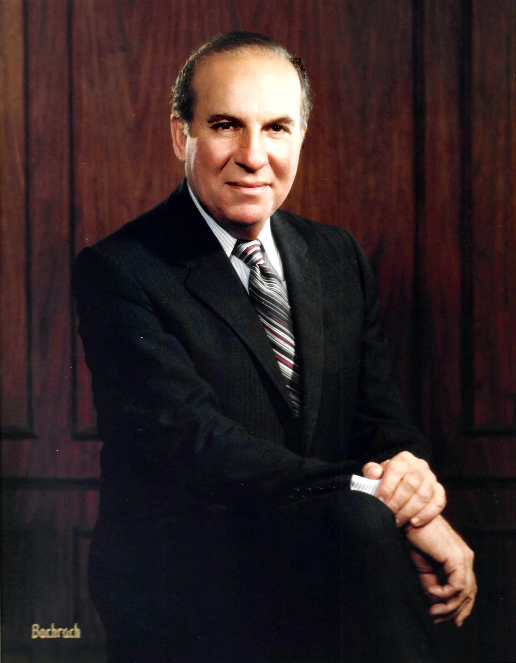 Portrait of a middle-aged man in a formal suit, smiling confidently against a wood-paneled background.